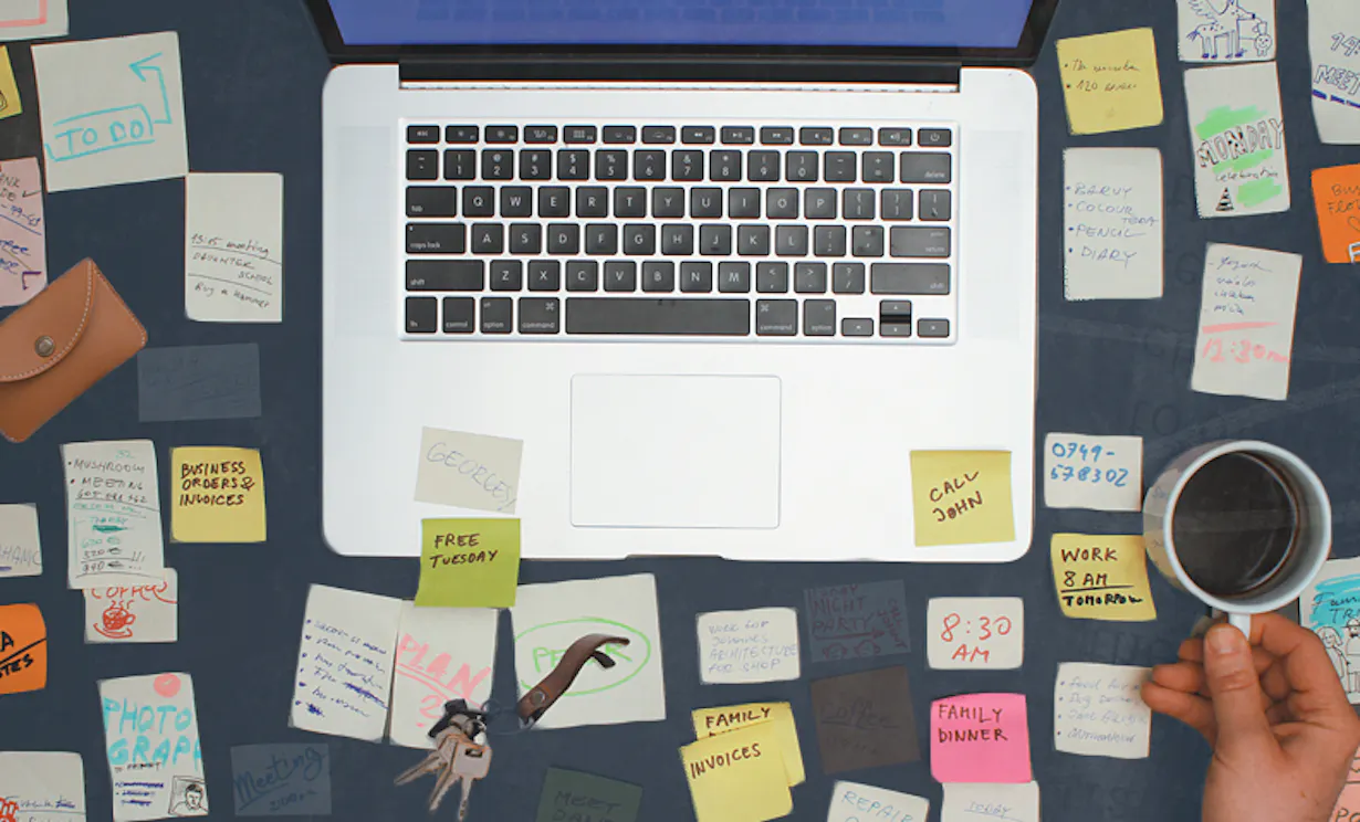 An overhead view of a desk with a computer open. The desk is filled with sticky notes and other objects showing reality of remote work.