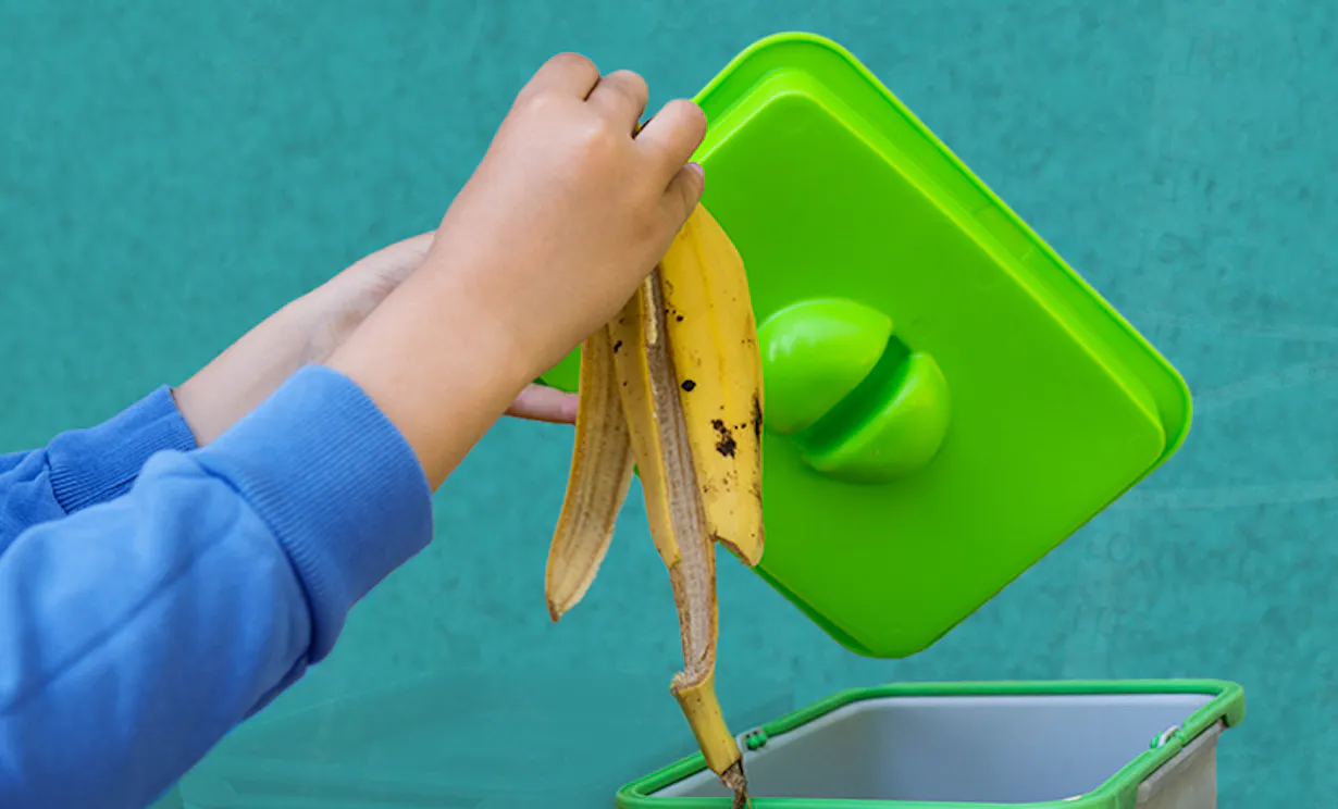 A hand placing a banana peel into an indoor compost bin