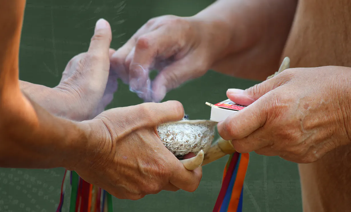 A person holding out their hands holding a shell with sage in it. The other holds a box of matches and lights the sage to perform a ceremonial smudge.
