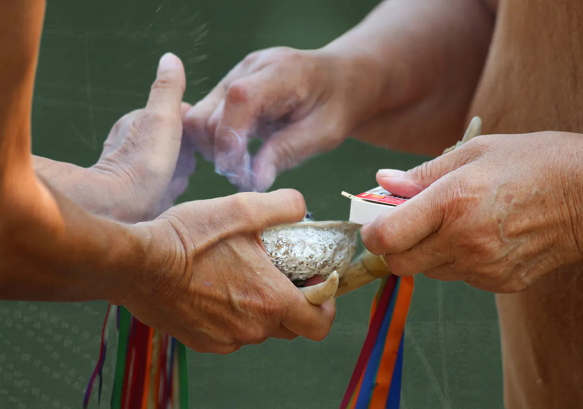 A person holding out their hands holding a shell with sage in it. The other holds a box of matches and lights the sage to perform a ceremonial smudge.