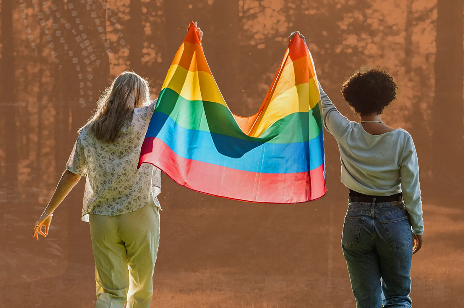 Two people hold a pride flag above their heads walking in nature