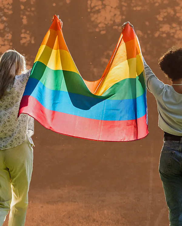 Two people hold a pride flag above their heads walking in nature