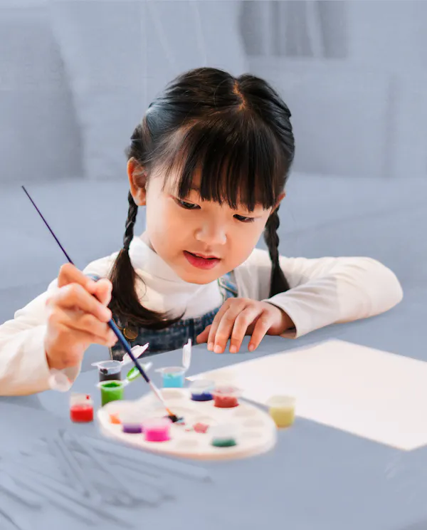 A small child sits at a coffee table painting on a piece of paper.