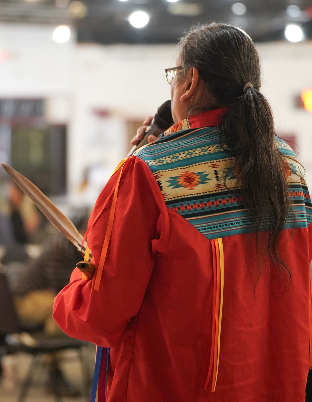 Back of man wearing a ribbon shirt while talking into a microphone with a feather in hand
