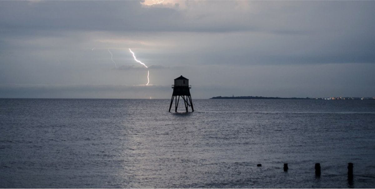 Lightning strikes in the sky behind lighthouse tower standing off shore