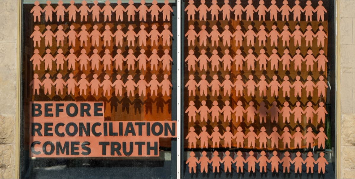Many orange paper children in a window display with "Before Reconciliation Comes Truth" written on the window