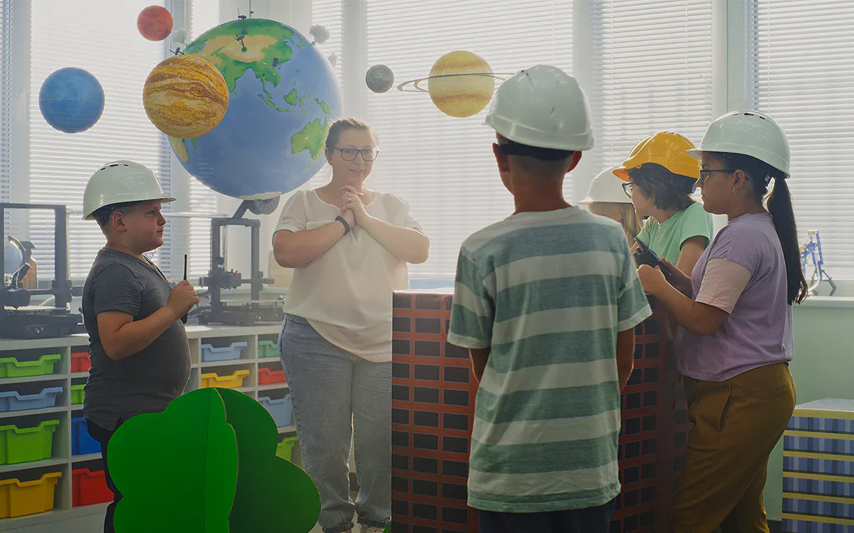 Children standing in a classroom surrounded by paper planets and coloured organizers. The kids are wearing hard hats and listening to a teacher.