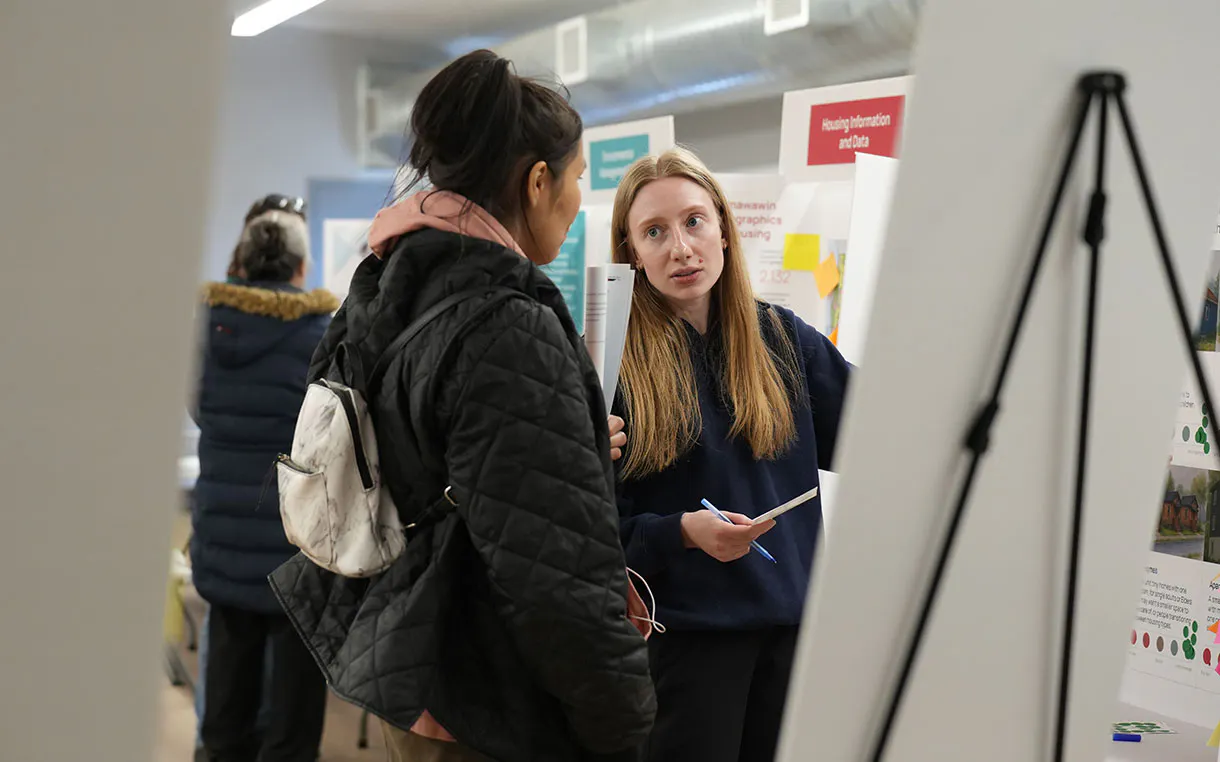 Two women speak to each other in during a community engagement. There are project poster boards throughout the room.