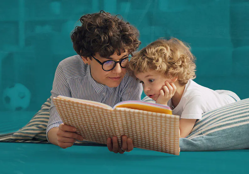 Parent and a young child are laying down on pillows and reading a book.