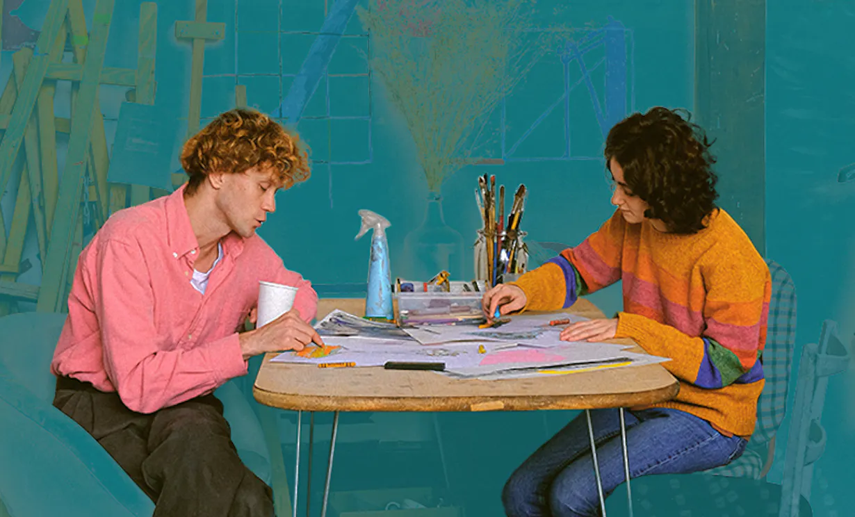 Two people sitting at a table drawing on paper with art supplies behind them.