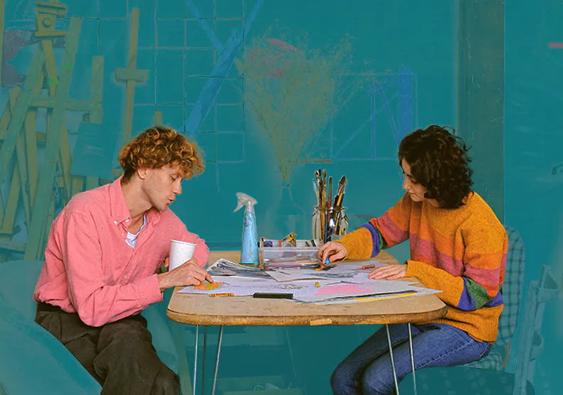 Two people sitting at a table drawing on paper with art supplies behind them.
