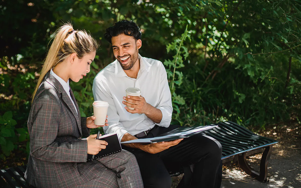 A man and woman sit on a park bench surrounded by greenery holding coffee cups and talking to each other