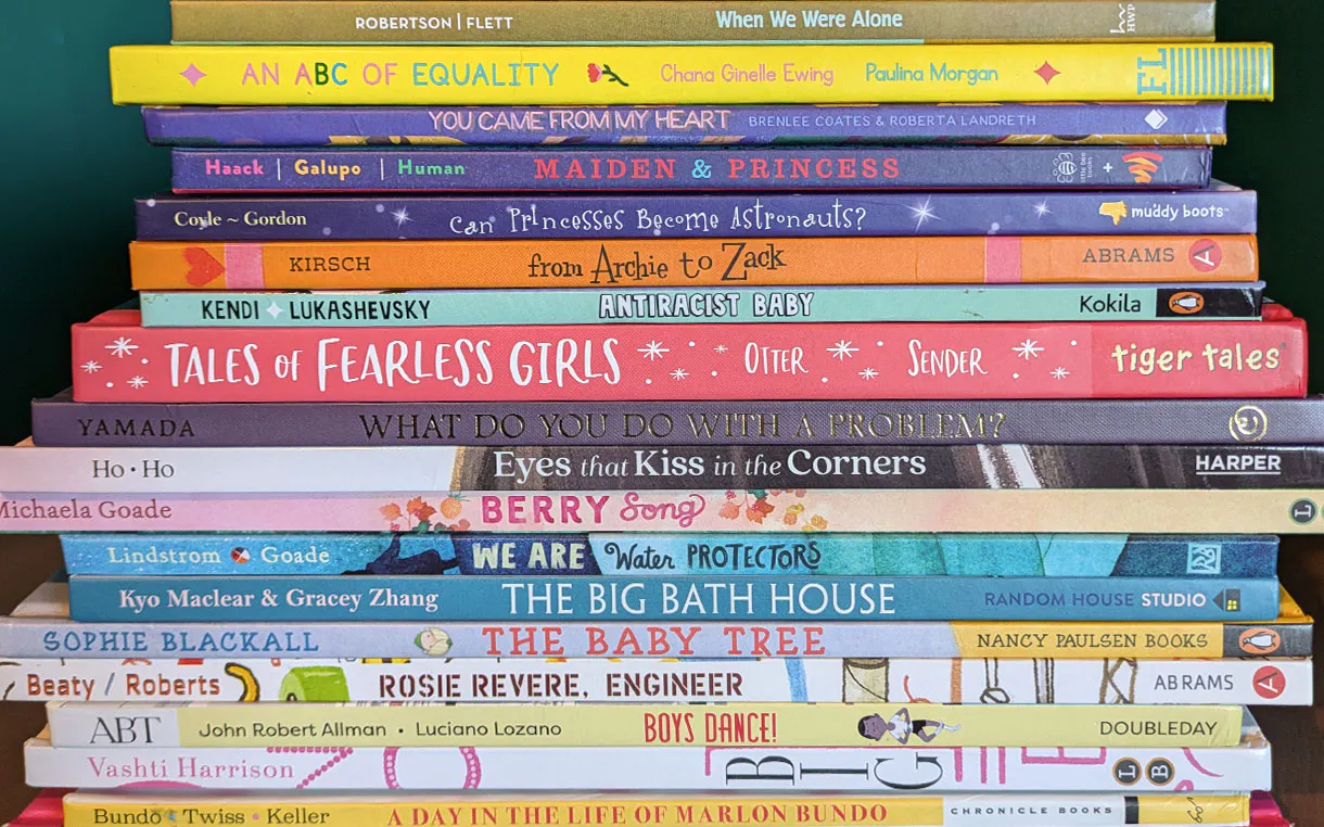 A tall, horizontal stack of colorful children’s picture books viewed from the side, showcasing book spines with diverse and inclusive titles. Notable titles include When We Were Alone, An ABC of Equality, Maiden & Princess, Antiracist Baby, Tales of Fearless Girls, Eyes that Kiss in the Corners, We Are Water Protectors, The Big Bath House, Rosie Revere, Engineer, Boys Dance!, Big, and A Day in the Life of Marlon Bundo.