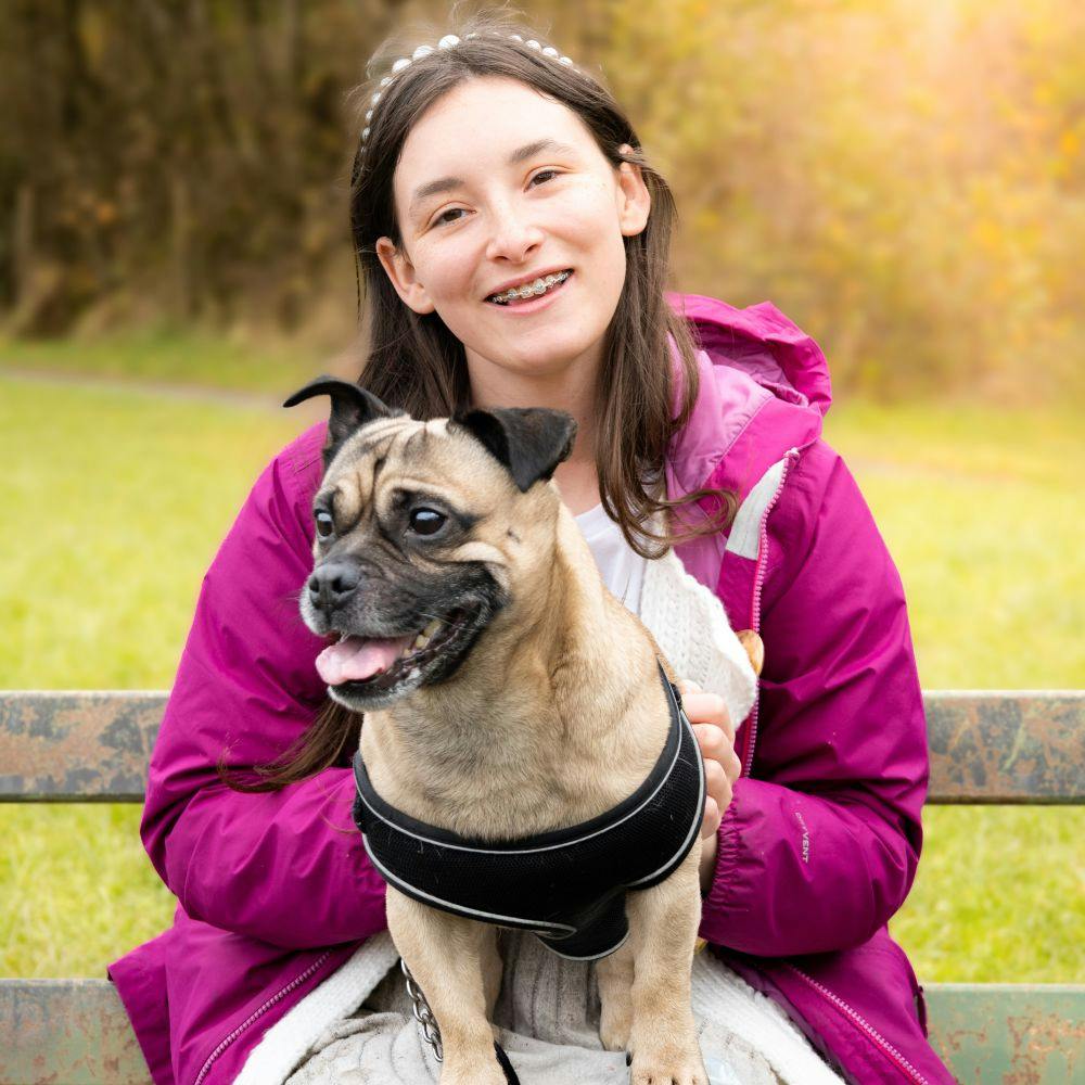 A smiling person wearing a pink jacket sits on a park bench, holding a small dog.