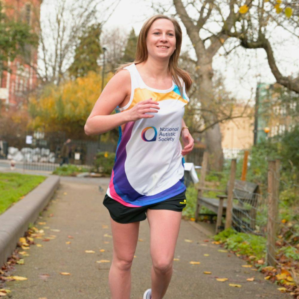 A smiling person runs along a park path, wearing a colourful National Autistic Society running vest.