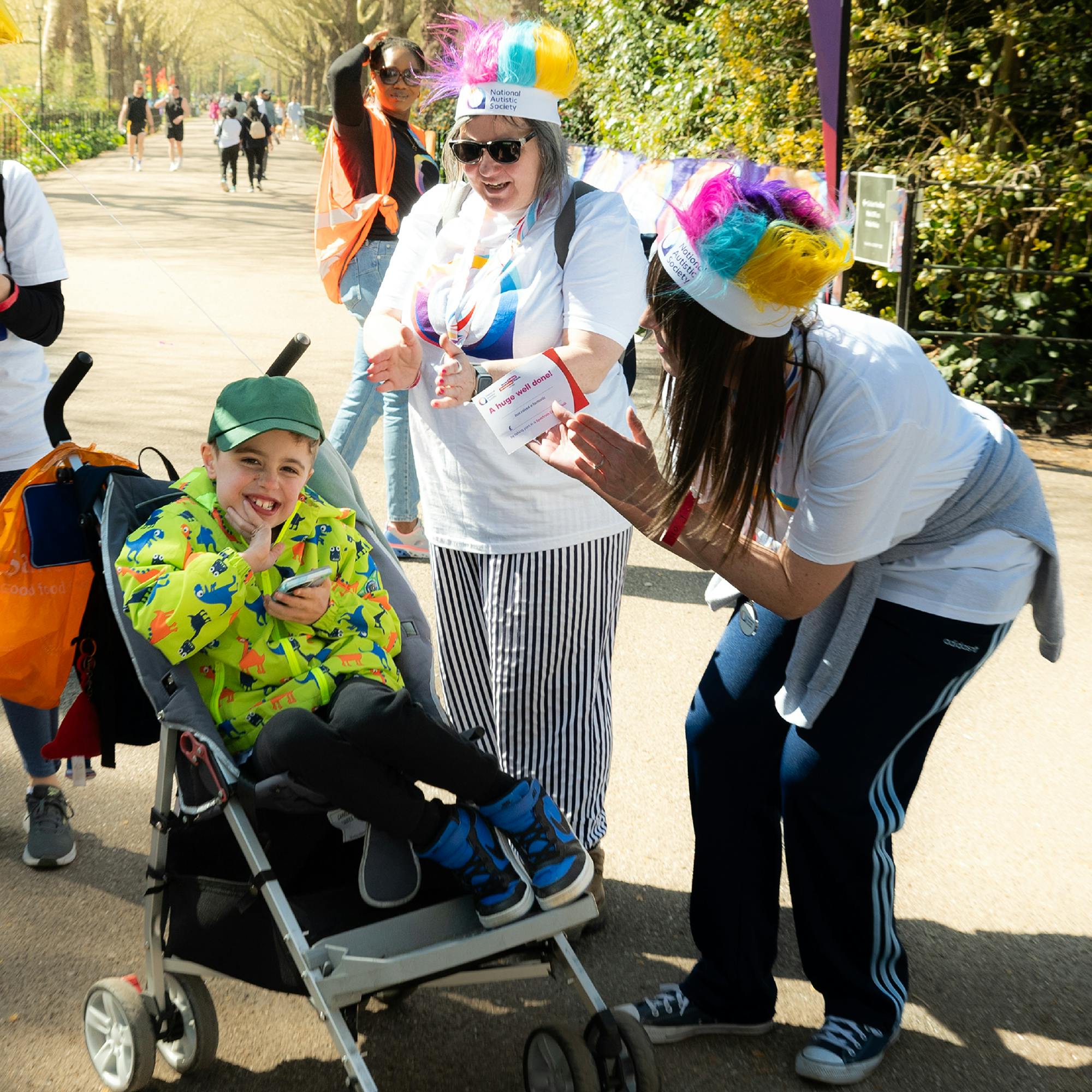 A smiling child in a bright yellow dinosaur jacket and green cap sits in a stroller, holding a phone, surrounded by two cheerful adults clapping and encouraging them. The adults are wearing white T-shirts with the National Autistic Society logo in blue, purple, and orange at an autism awareness event on a sunny day.