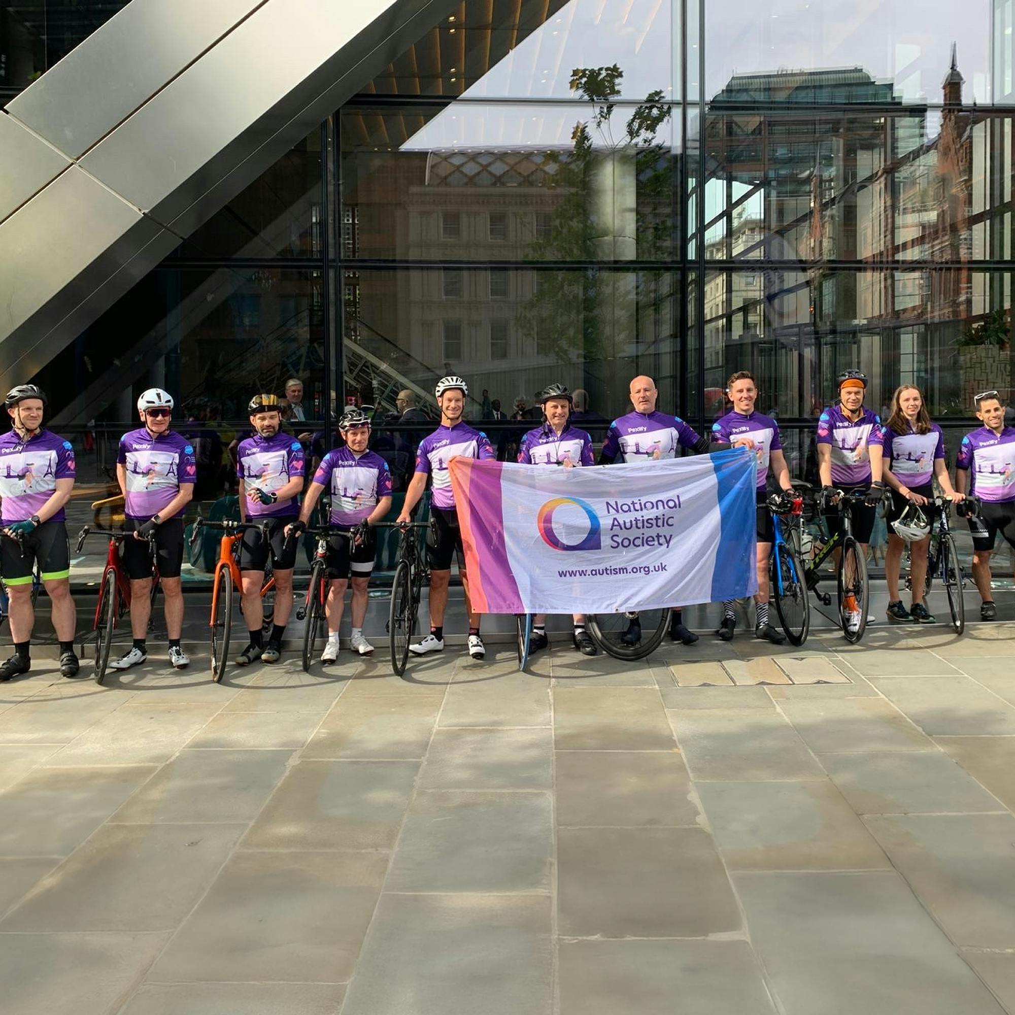 A group of cyclists standing in a row with their bicycles in front of a modern glass building. They are wearing matching purple cycling jerseys and holding a large banner that reads “National Autistic Society” with the website www.autism.org.uk and a colorful circular logo.