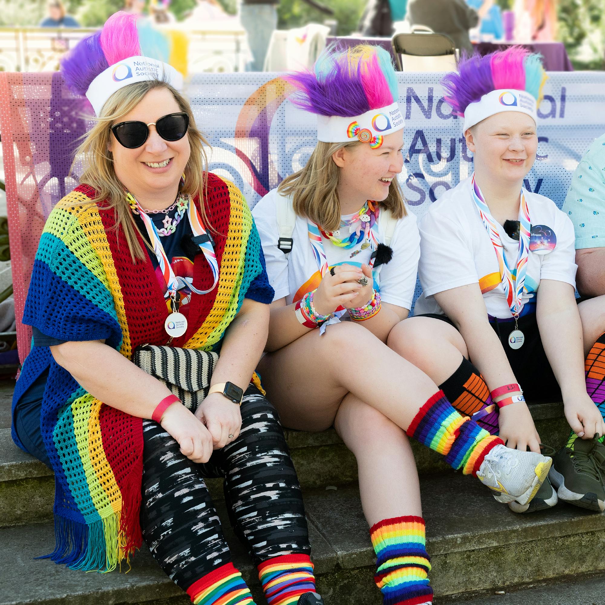 Three people sitting outdoors on stone steps wearing colorful outfits and accessories for an autism awareness event. They have rainbow-themed clothing, including striped socks, patterned leggings, and a crocheted rainbow shawl. Each person wears a white headband with a purple and pink mohawk-style decoration and a lanyard with badges. Behind them is a banner displaying the National Autistic Society logo.