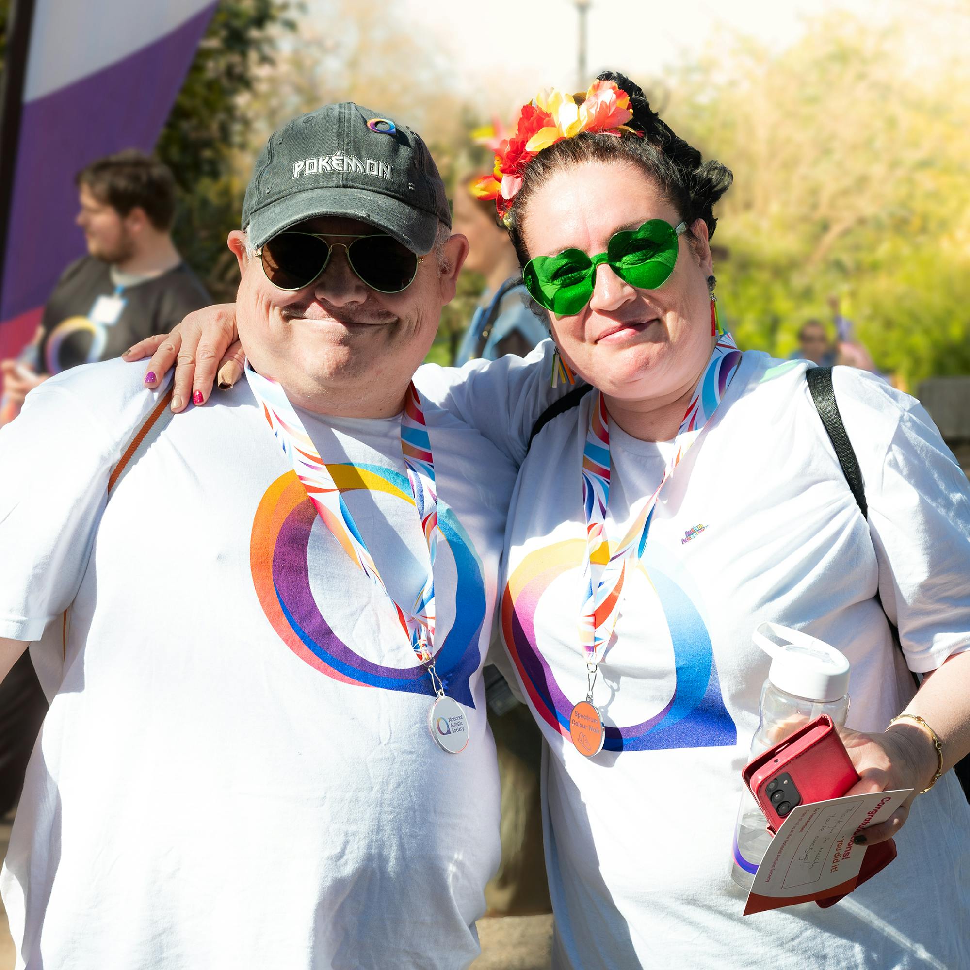Two people standing close together outdoors at an autism awareness event, wearing white T-shirts with the National Autistic Society logo in blue, purple, and orange. Both have striped lanyards with medals around their necks. One person is wearing a dark cap with “Pokémon” text, and the other has a colorful flower headband and is holding a water bottle and a red phone case. Trees and other participants are visible in the background.