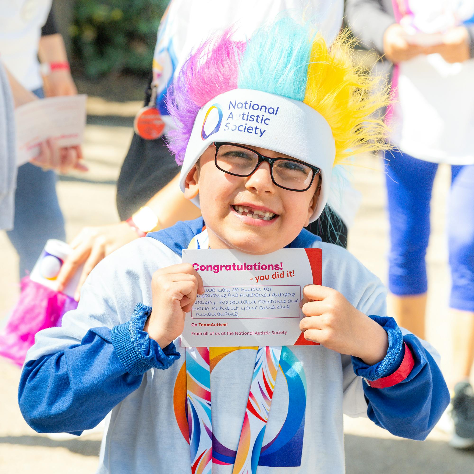 Small boy wearing a white headband with a colorful mohawk in blue, pink, and yellow, along with a National Autistic Society T-shirt and a rainbow-striped lanyard. The boy is holding a card that reads “Congratulations! You did it!” in bold red text.