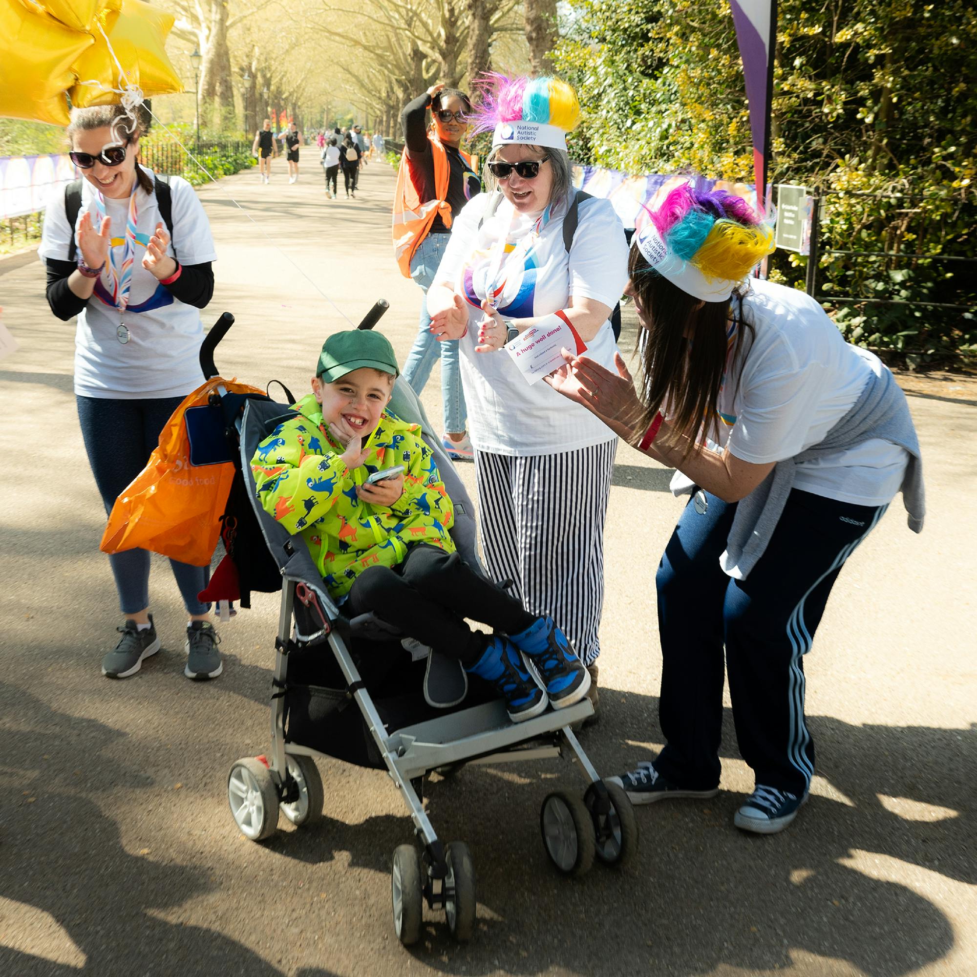 A smiling child in a bright yellow dinosaur jacket and green cap sits in a stroller, holding a phone, surrounded by three cheerful adults clapping and encouraging them. The adults are wearing white T-shirts with the National Autistic Society logo in blue, purple, and orange at an autism awareness event on a sunny day.