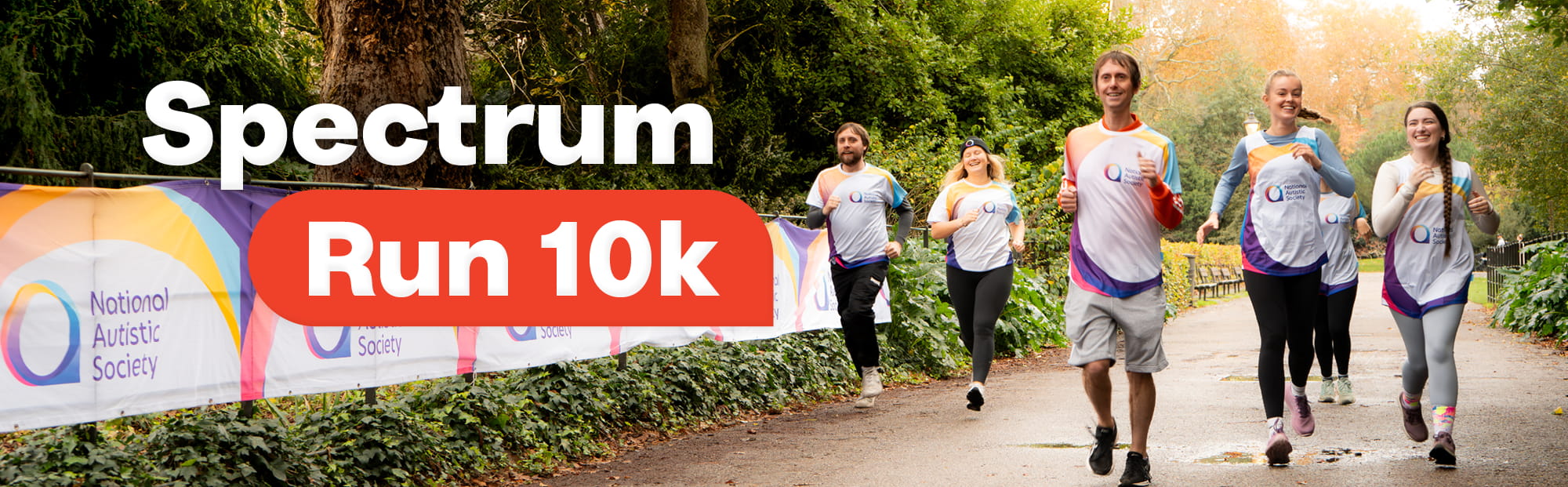 A group of smiling runners wearing National Autistic Society–branded shirts jog along a tree-lined park path. A large banner on the left displays the charity’s logo and colourful design. Bold text on the image reads 'Spectrum Run 10k.'