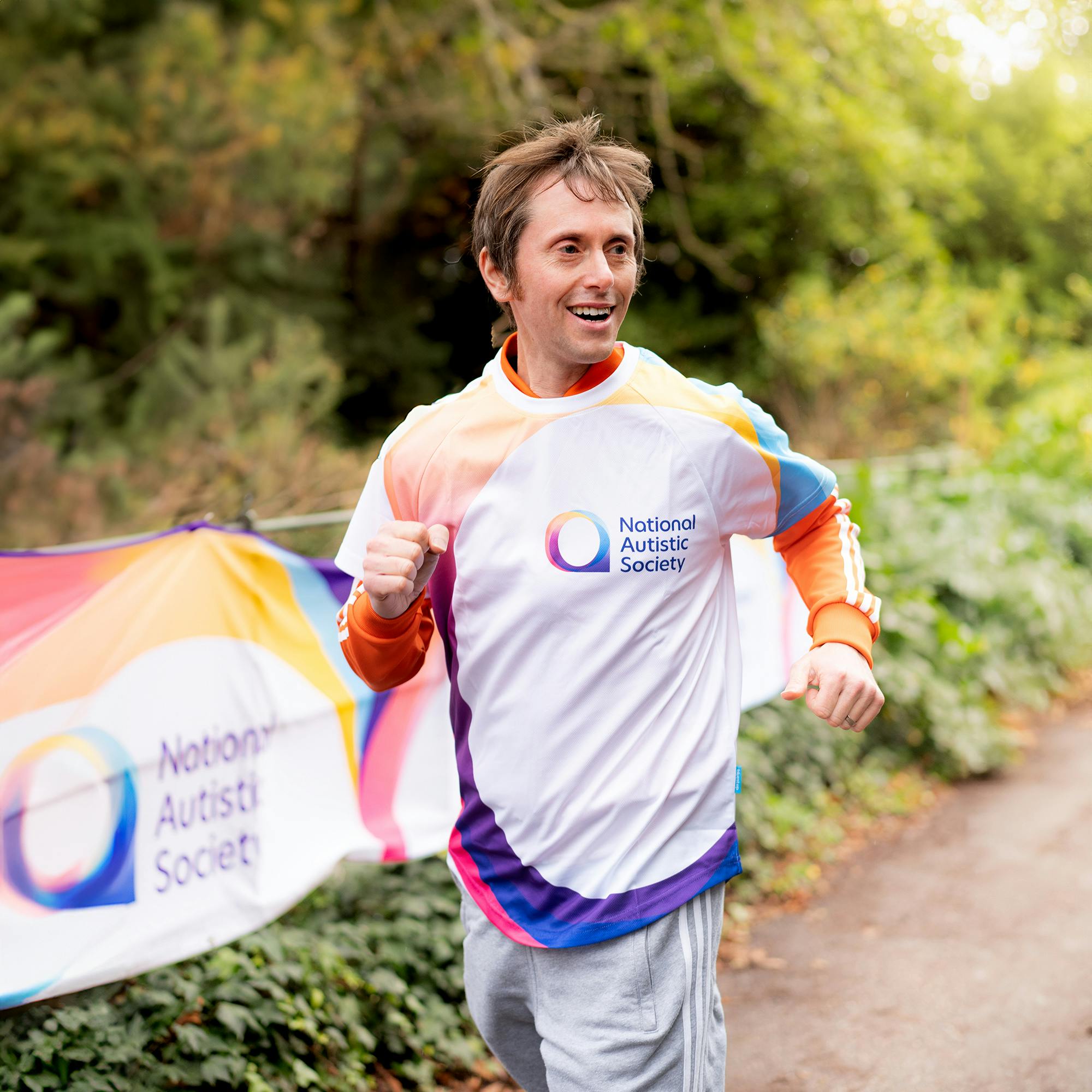 Person running outdoors wearing a white long-sleeve top with colourful accents and the National Autistic Society logo. A matching banner with the same logo and colours is visible along the path.