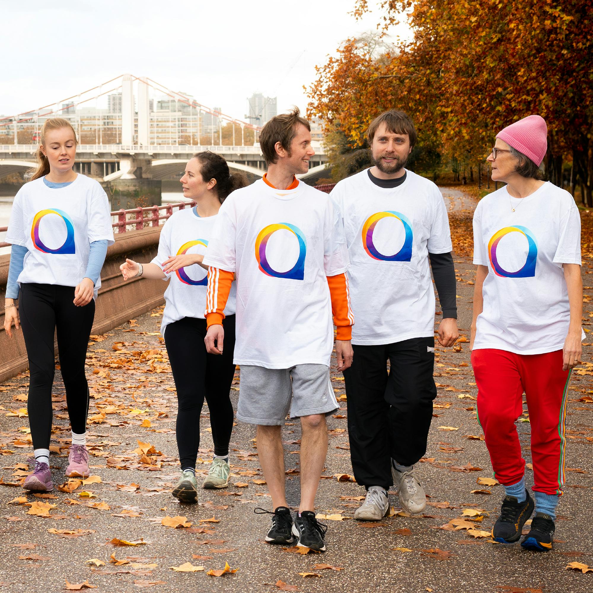 A group of adults wearing National Autistic Society T-shirts walk together along a riverside path lined with autumn leaves. A bridge and city buildings appear in the background. 