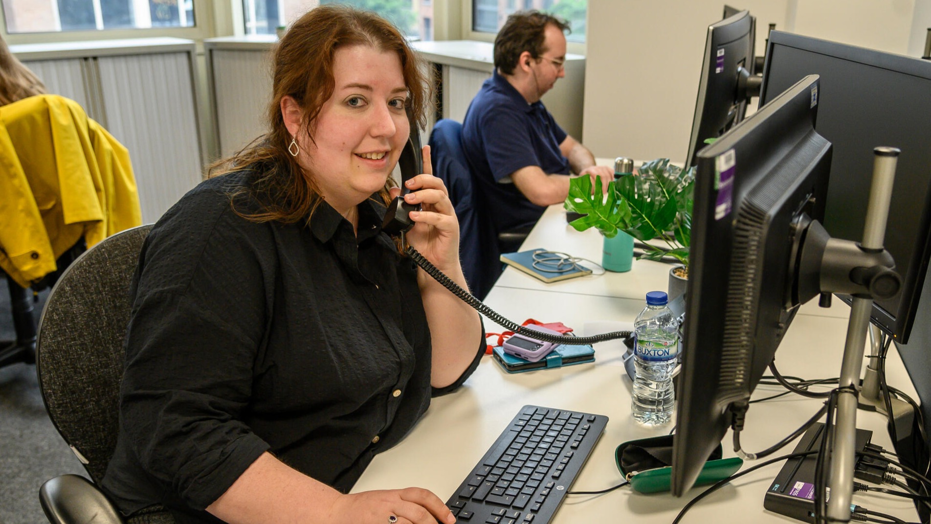 Person sitting at an office desk using a landline phone while typing on a keyboard. The desk has a computer monitor, a water bottle, a smartphone, and a small green plant. Another person is seated at a desk in the background, also working on a computer. Large windows are visible behind them.