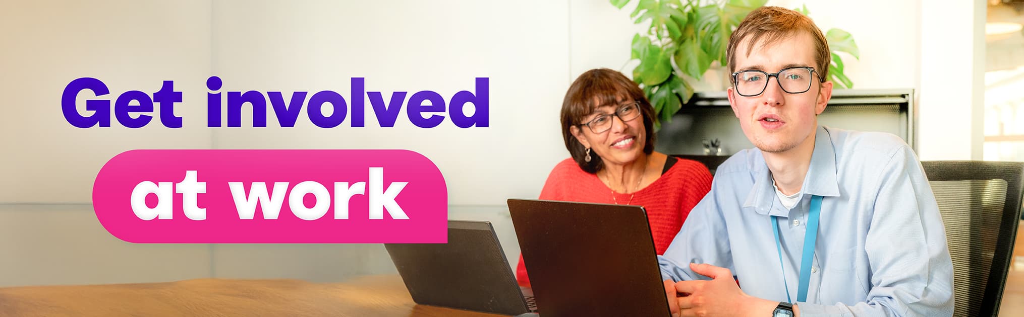 Two people sitting at a table with laptops in an office setting. A green plant is visible in the background. Large text on the left side reads “Get involved” in purple and “at work” in white inside a pink rounded rectangle.