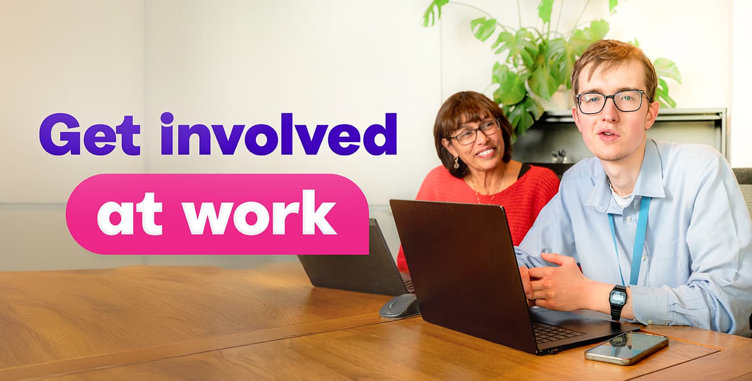 Two people sitting at a table with laptops in an office setting. A green plant is visible in the background. Large text on the left side reads “Get involved” in purple and “at work” in white inside a pink rounded rectangle.