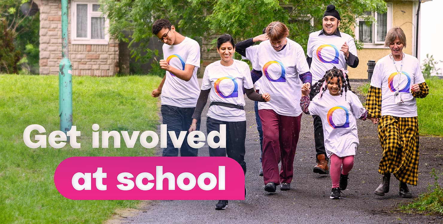 A group of six people walking outdoors on a paved path, all wearing matching white T-shirts with a colorful circular logo. In the background, there are green trees, grass, and a building. On the left side of the image, large text reads 'Get involved at school'.