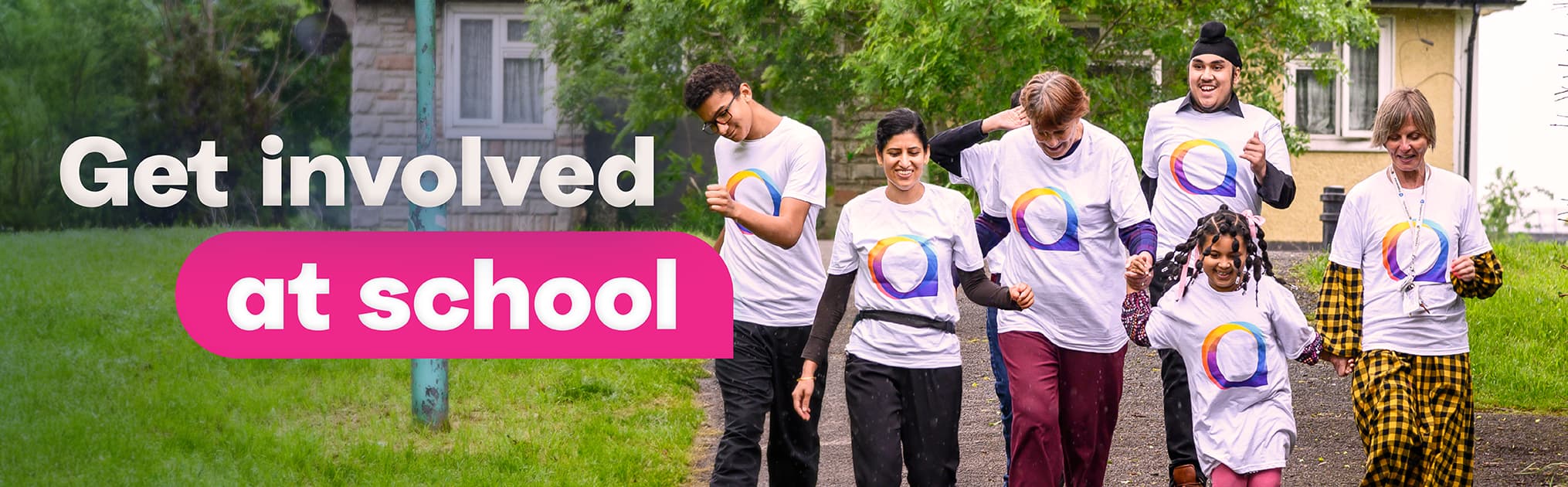 A group of six people walking outdoors on a paved path, all wearing matching white T-shirts with a colorful circular logo. In the background, there are green trees, grass, and a building. On the left side of the image, large text reads 'Get involved at school'.