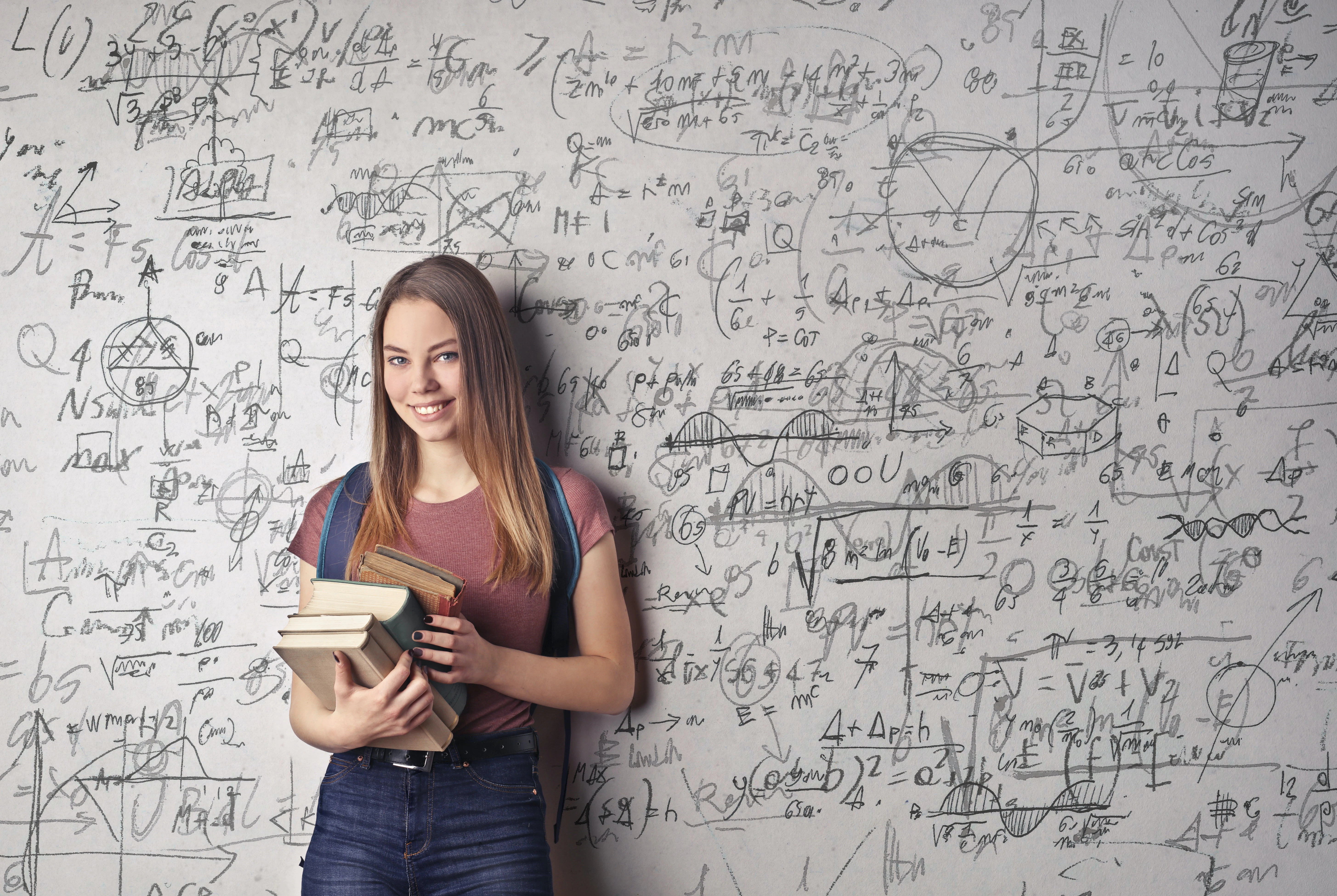 A college student with brown hair, holding a stack of books, standing in front of a white board with math written on it