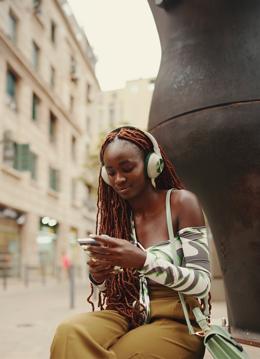 Young woman listening to headphones, smiling. She is sitting on a wall, outdoors.