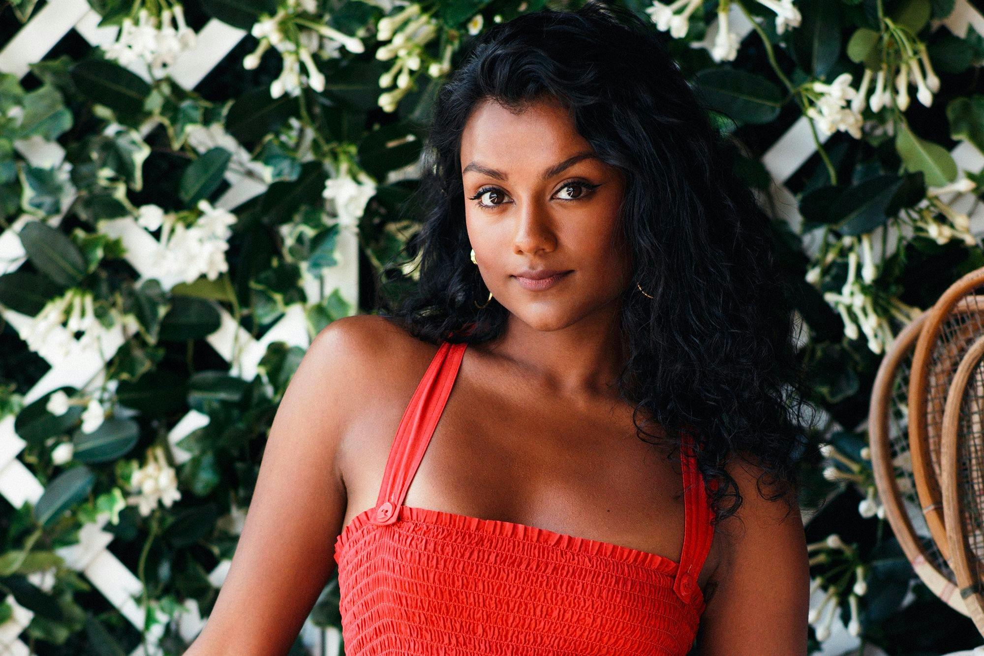 Simone Ashley wears a red dress against a flower covered trellis. Her hair is dark and long, and smiles slightly.