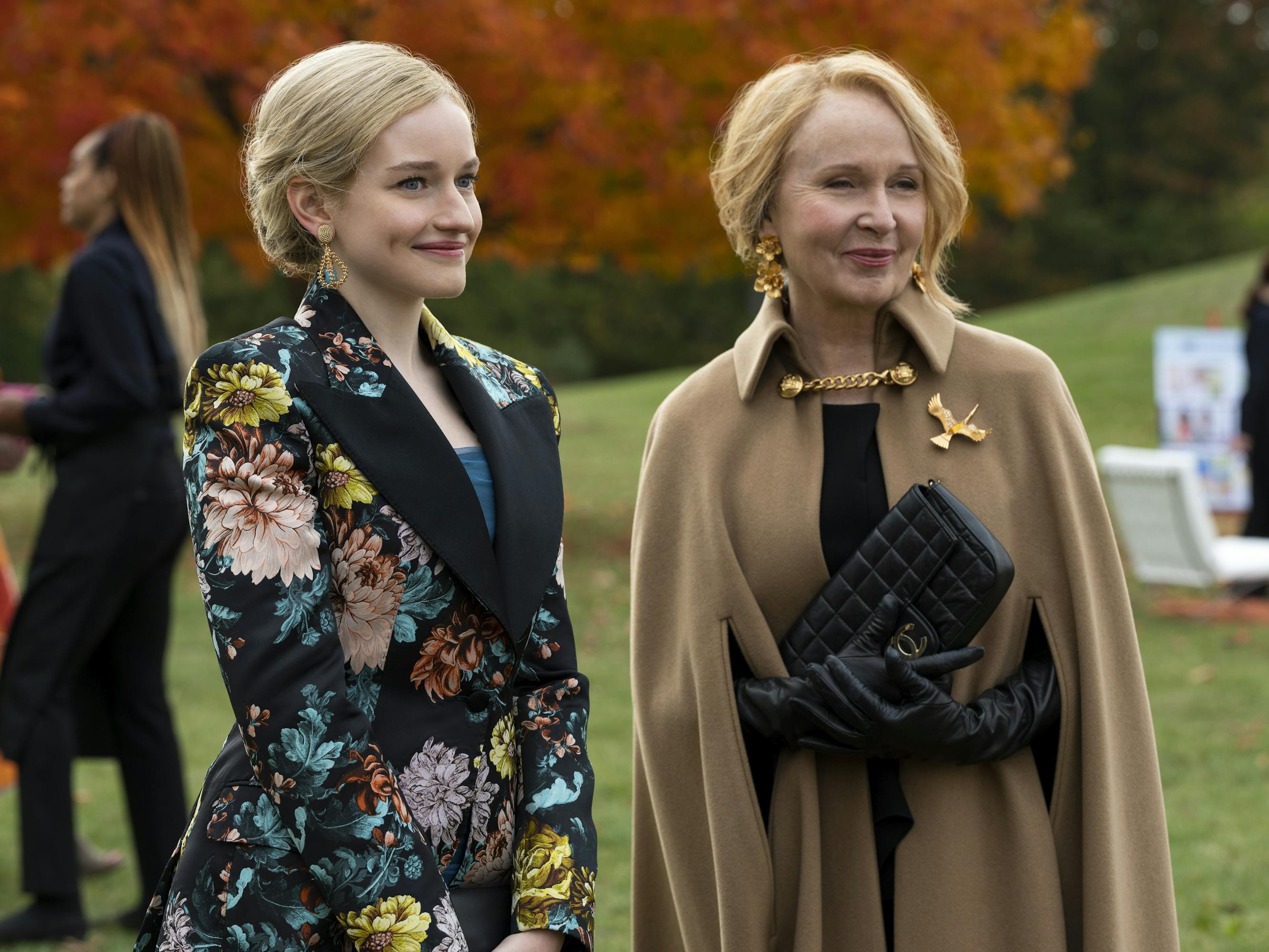 Anna Delvey and Kate Burton stand together outside. Delvey wears a flowery jacket. Burton wears a camel coat, black gloves, and a black clutch.