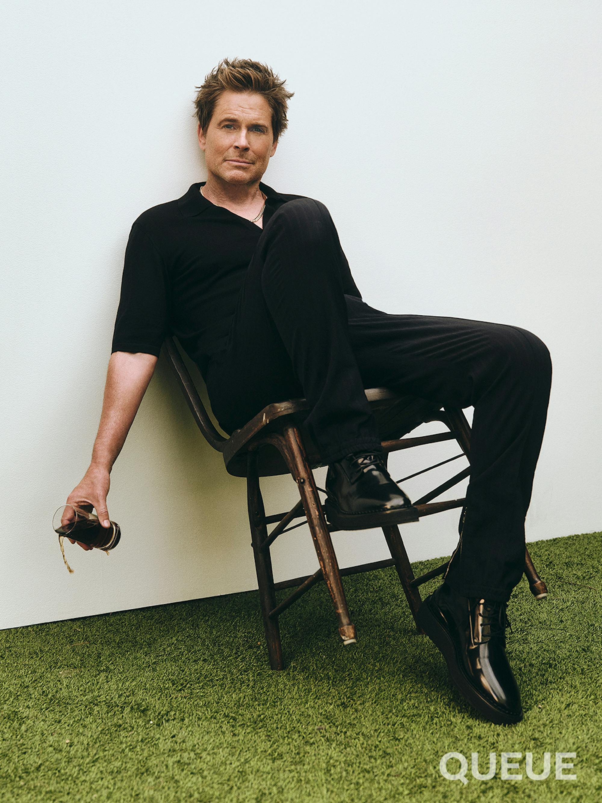 Rob Lowe wears all black, including black dress shoes, and pours some cold brew on the AstroTurf ground as he leans back in his chair. A very precarious sight!