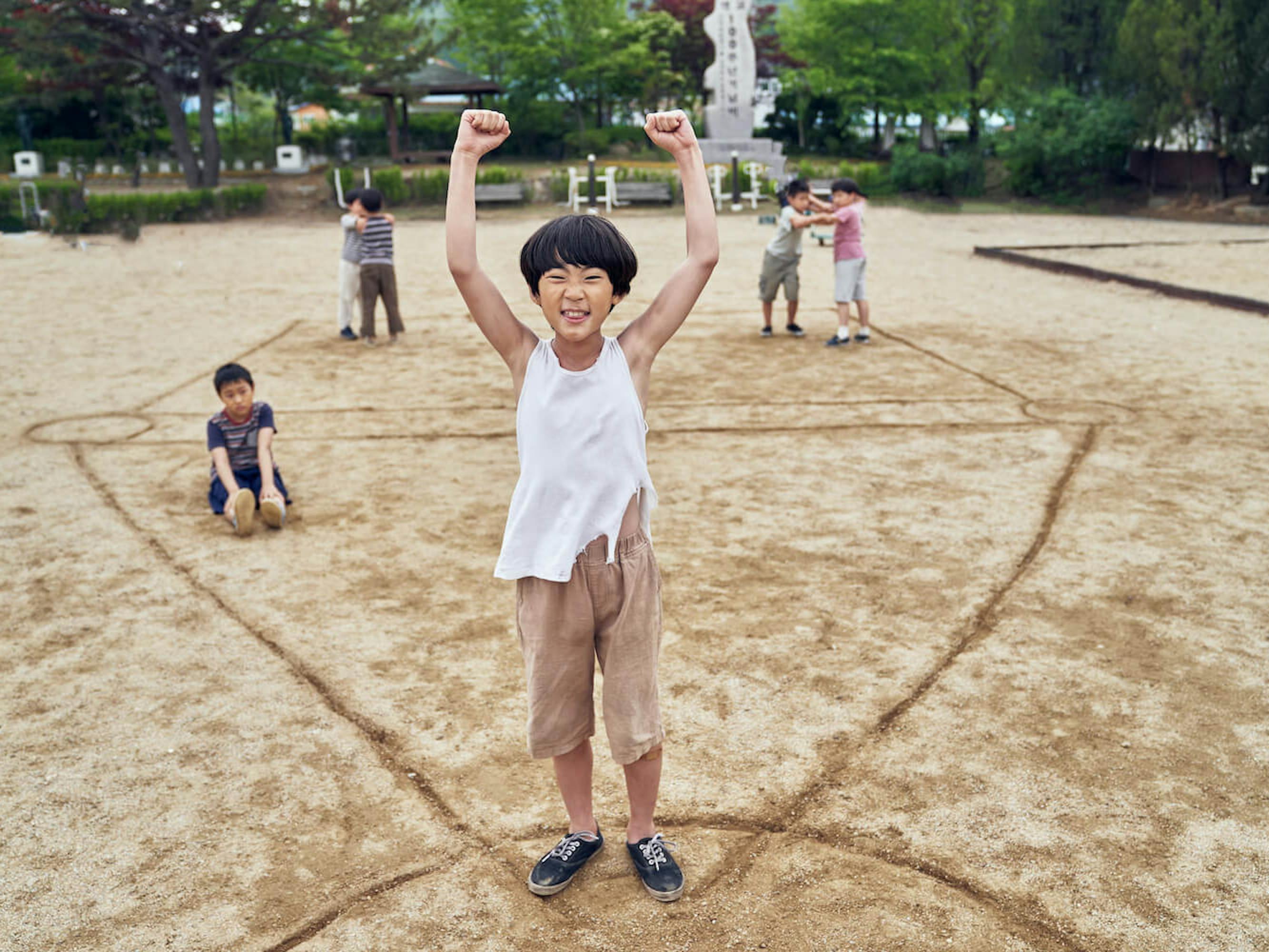 A young child raises their hands wearing khakis and a white tank top. Behind them are a bunch of little kids playing in the sand.
