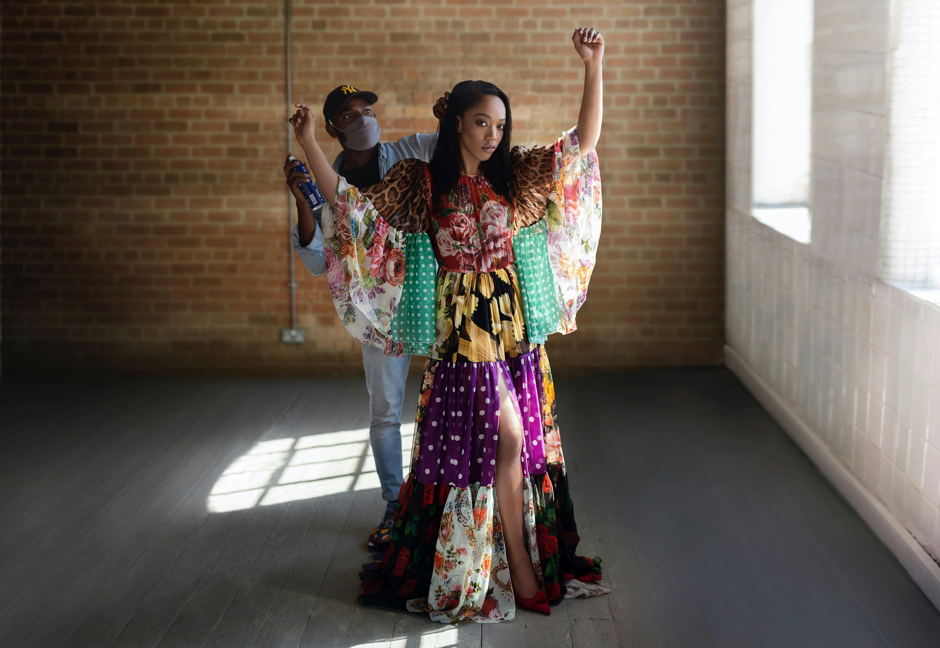 Naomi Ackie stands with her arms raised and wearing a colorful maxi dress of patchwork patterned fabrics. A hairstylist wearing a face mask stands behind Naomi, applying hairspray to her long dark hair.