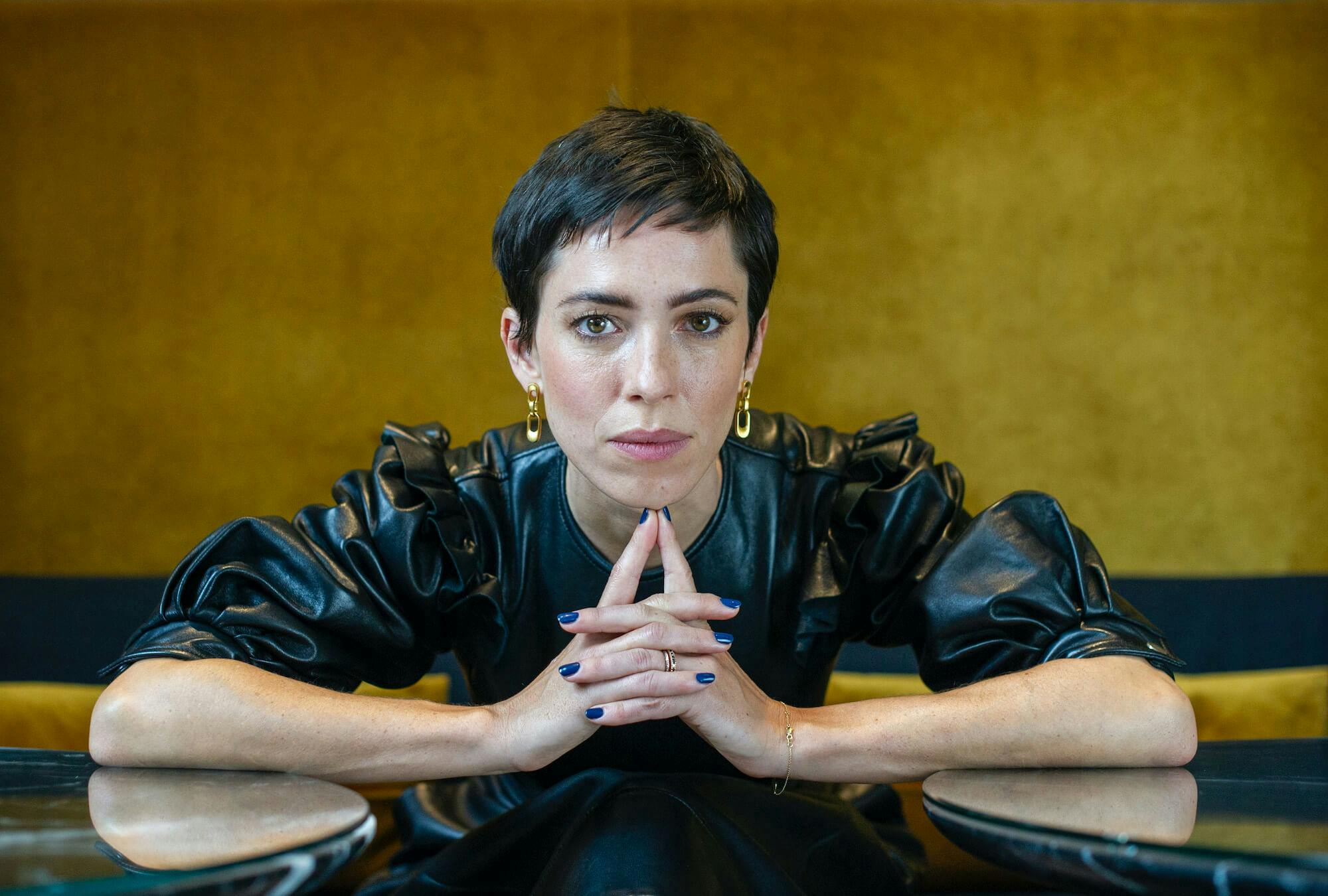 Rebecca Hall gazes into the camera, her arms resting on the table and hands clasped in front.  She wears a black ruffled leather dress 