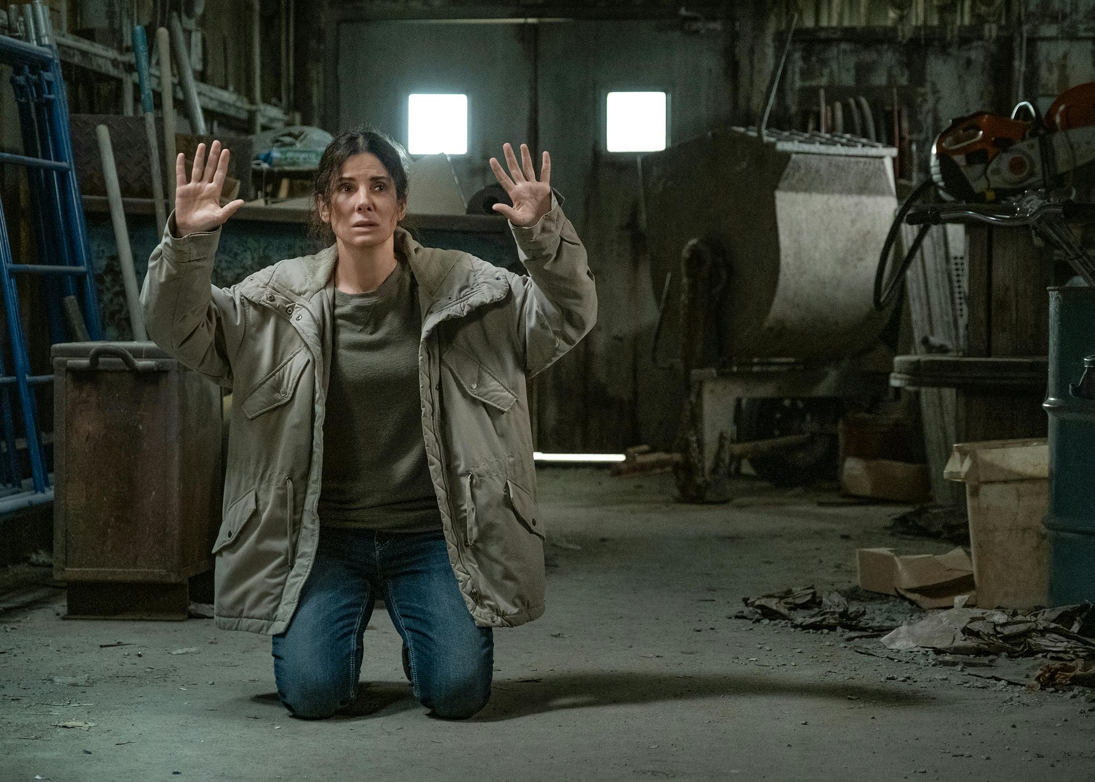 Ruth Slater (Sandra Bullock) kneels in a dusty room. She wears blue jeans, an olive shirt, and light green jacket. 