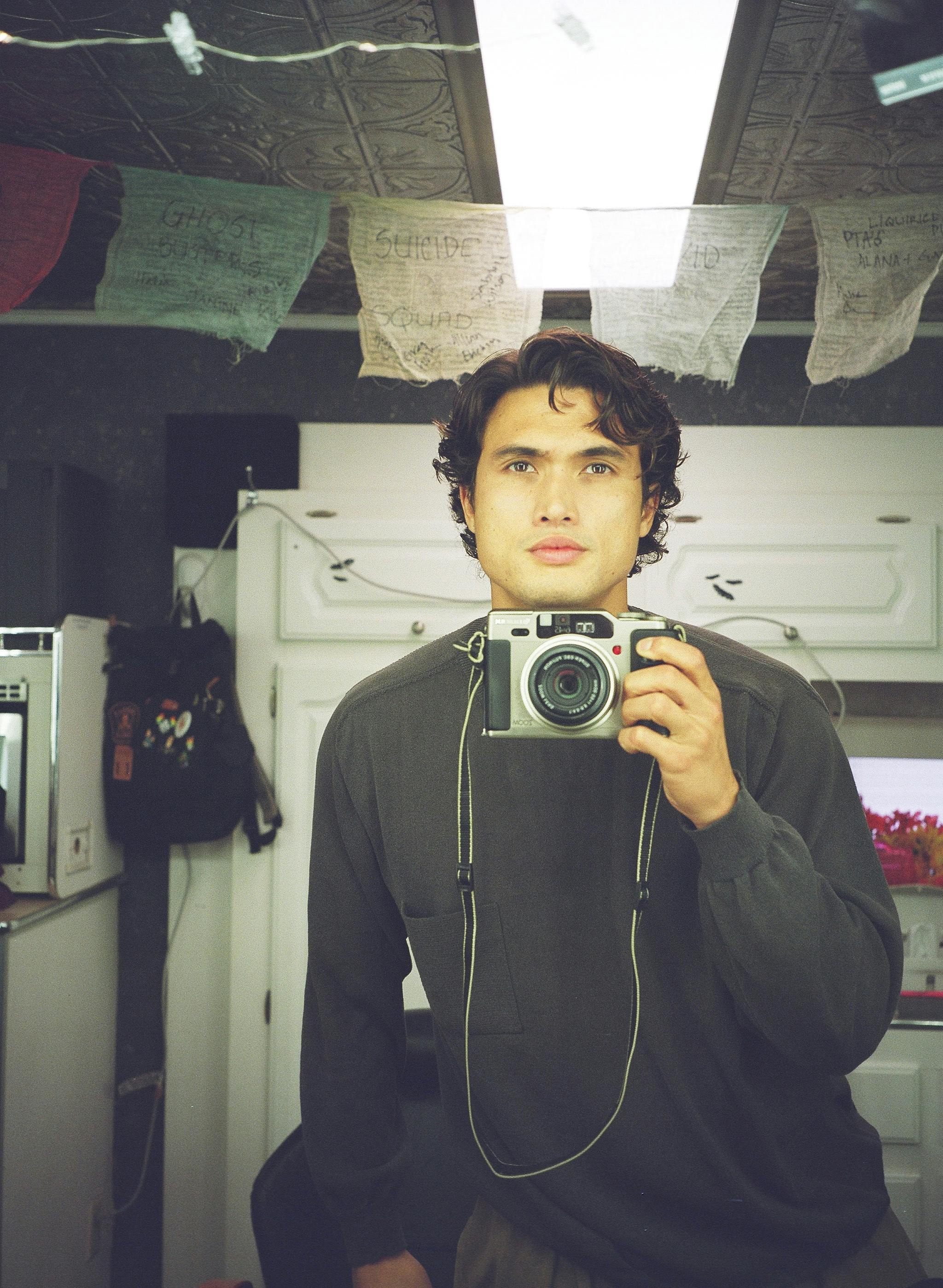 Charles Melton smizes and takes a selfie with a film camera. 