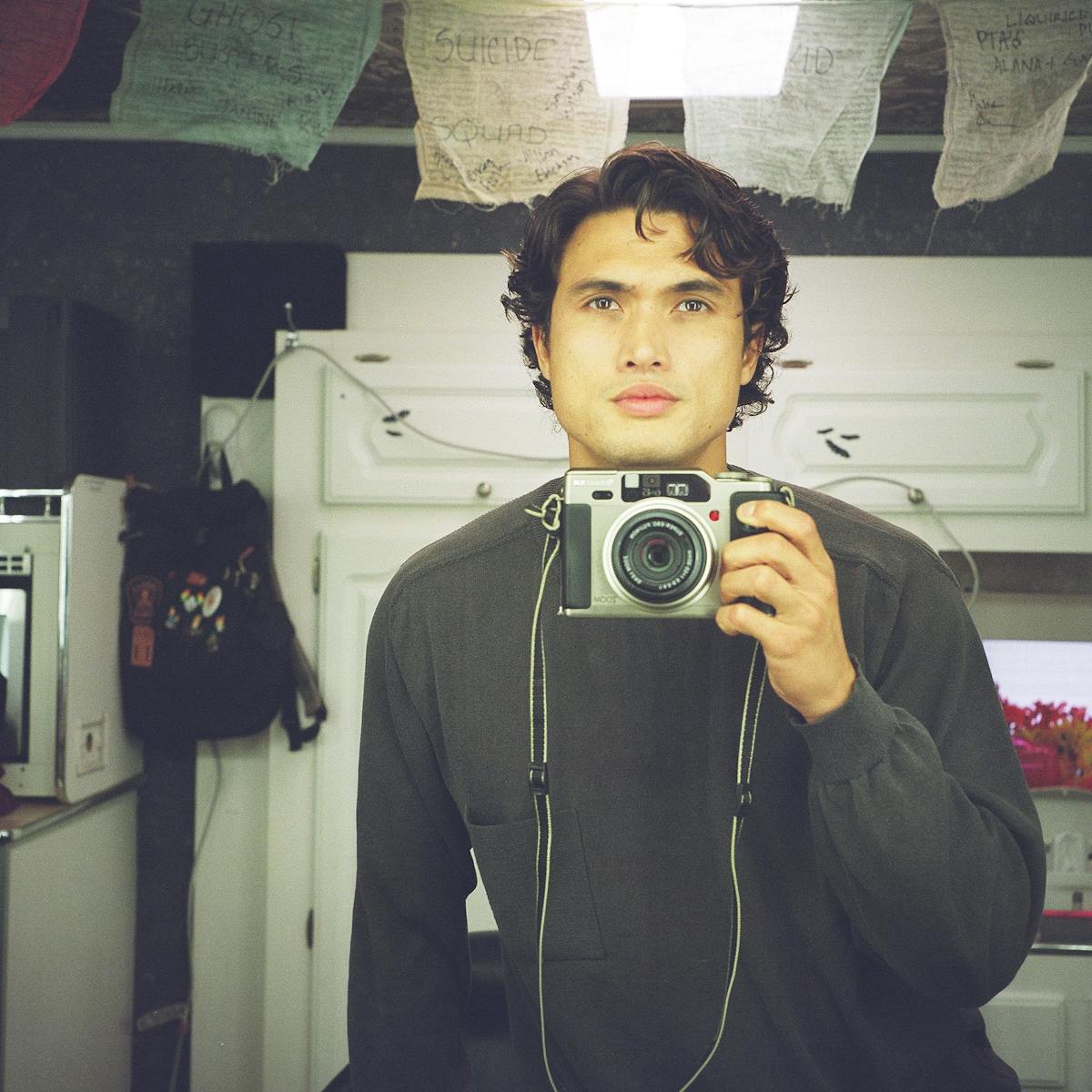 Charles Melton smizes and takes a selfie with a film camera. 