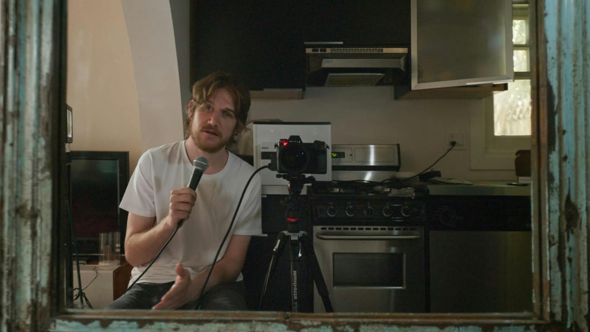 Bo Burnham sings into a microphone looking in a mirror. He has camera equipment, and a kitchen in the background. The mirror is edged with a blue frame.
