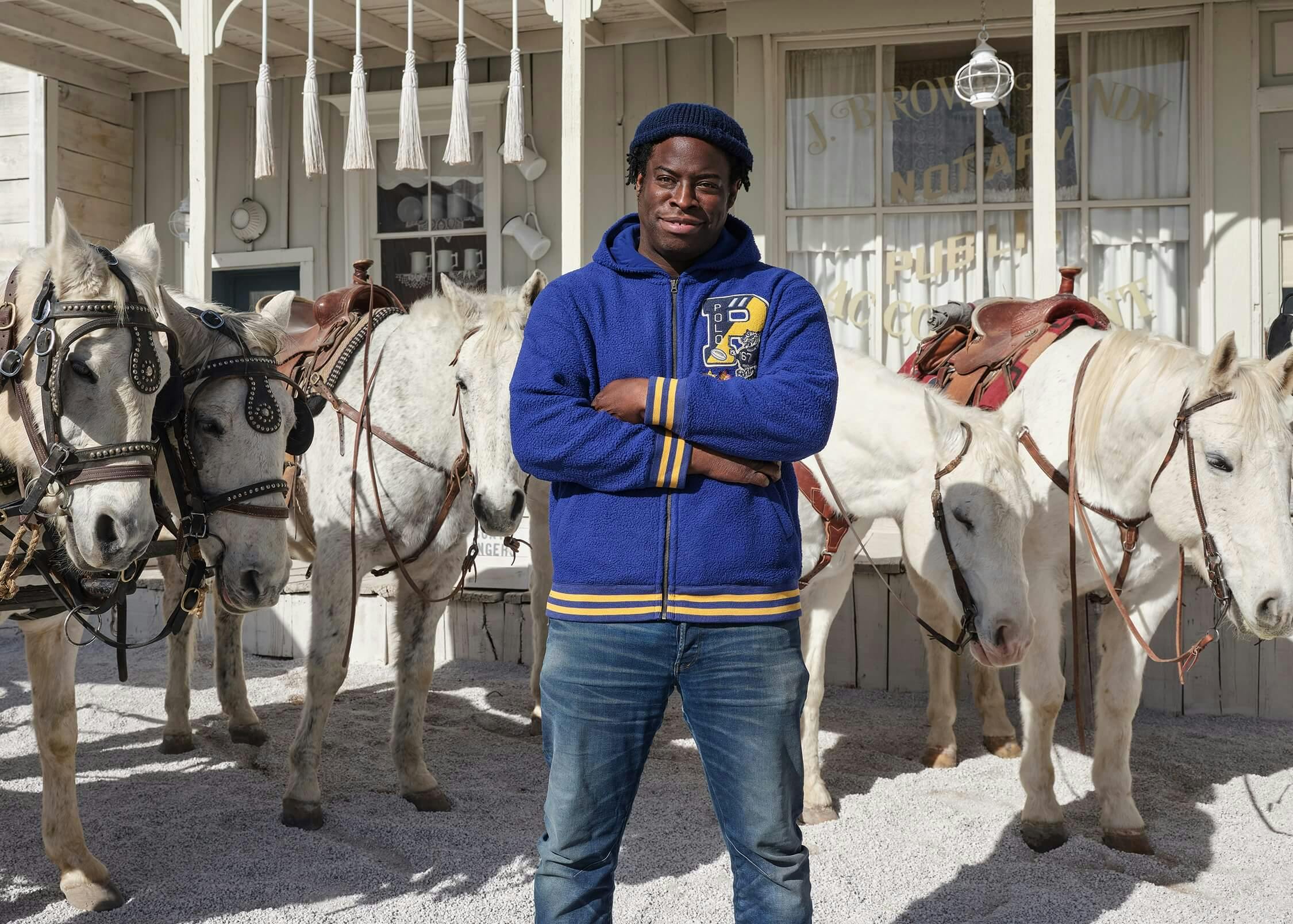 Jeymes Samuel wears a blue and yellow hoodie, navy hat, and blue jeans and stands with his arms crossed. Behind him are five white horses, and a white house.