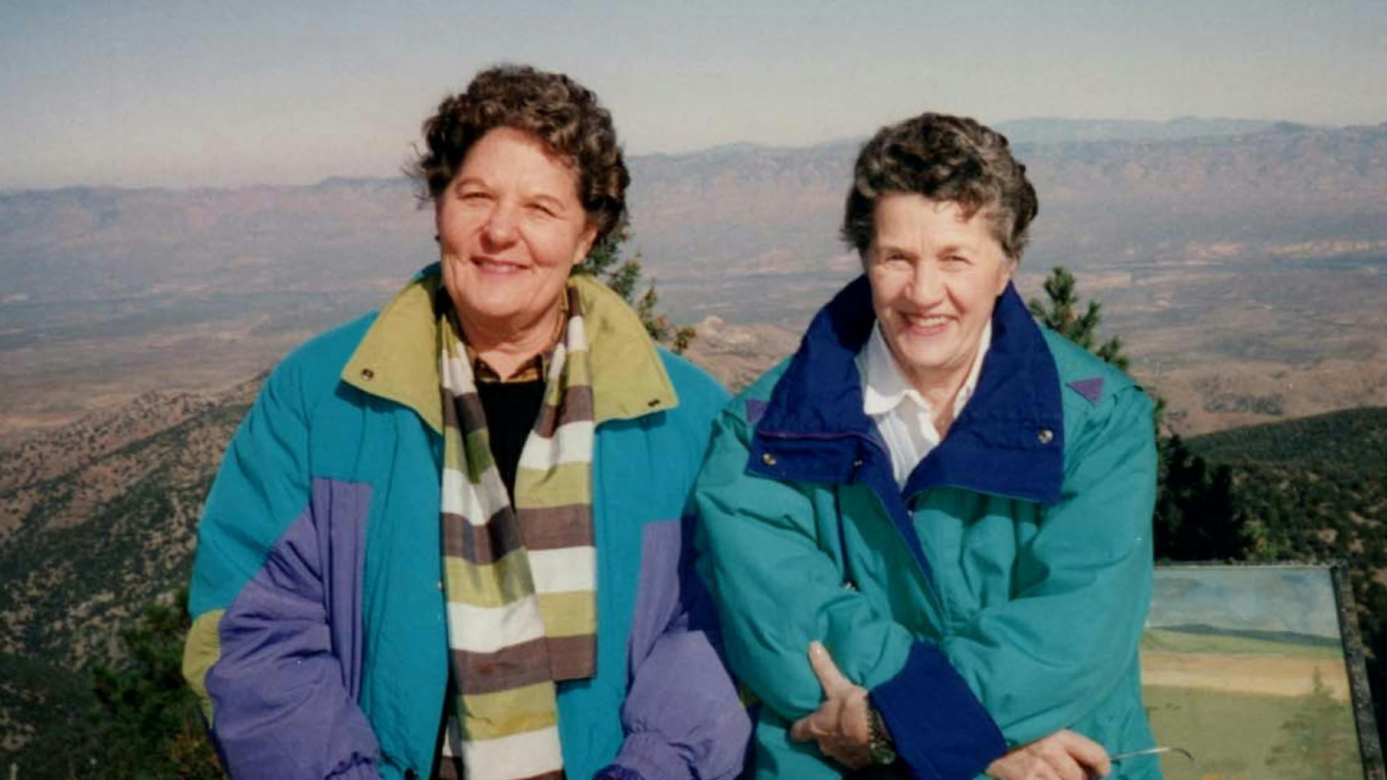Henschel and Donahue pose smiling in front of scenic mountains in this faded photograph. The pair wear windbreakers in classic 80s colors.  