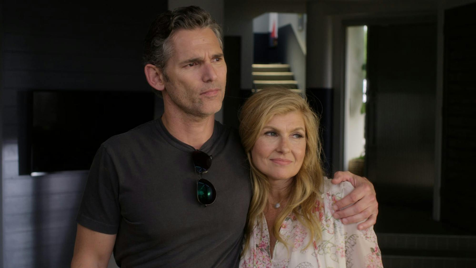 Connie Britton and Eric Bana stand together in Dirty John. He has his arm around her, and they stand in semi darkness.