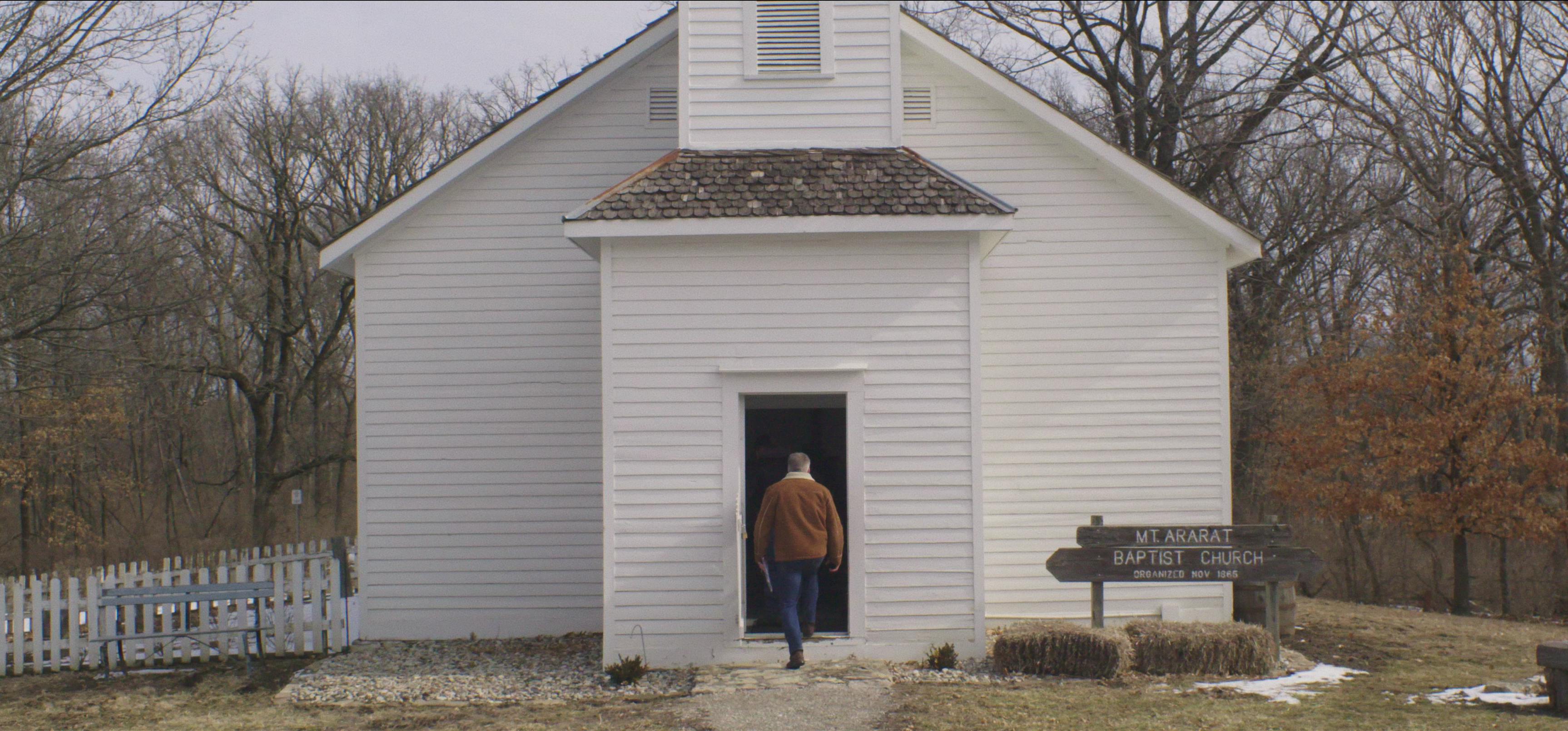 Dan Laurine enters a small white wooden church surrounded by bare trees and there are a couple of small patches of snow on the ground.