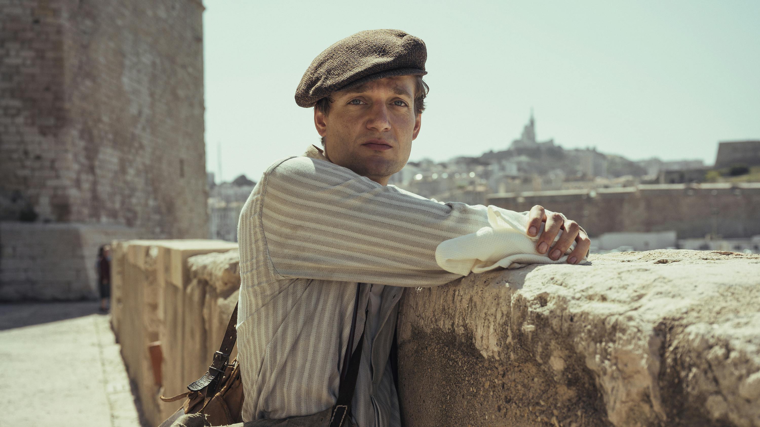 Albert Hirschman (Lucas Englander) wears a striped shirt and messenger boy hat and leans against a crumbly ledge.